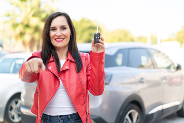 Middle aged woman holding car keys at outdoors points finger at you with a confident expression