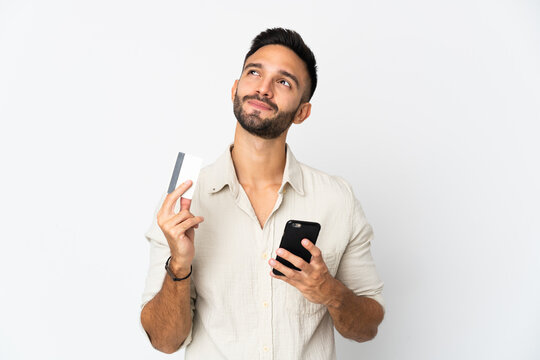 Young caucasian man isolated on white background buying with the mobile with a credit card while thinking