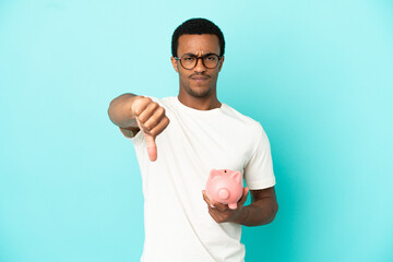 African American handsome man holding a piggybank over isolated blue background showing thumb down...