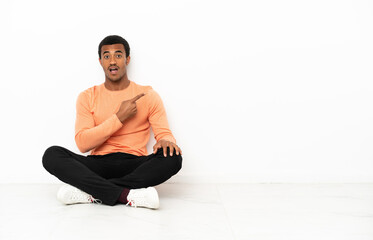 African American man sitting on the floor over isolated copyspace background surprised and pointing side