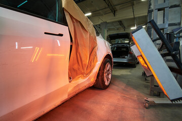 A white car is undergoing body repair with infrared drying equipment. The vehicle's side panel is masked with paper for protection during the heating process in an auto repair shop.