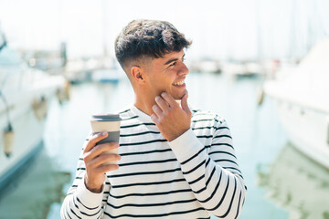 Young hispanic man holding a take away coffee at outdoors thinking an idea and looking side