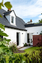 A white cottage in the Scottish Highlands, with a garden and a modern-style house with a black roof. The cottage has a gable window on one side and is directly connected to the main family home.