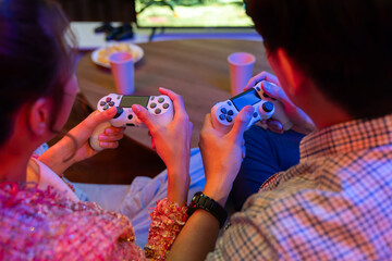 Naklejka premium Cropped of hands couple with joysticks sitting sofa playing video game together at back side view with fronted snack and drinks with fun mood at neon light color living room at modern home. Infobahn.