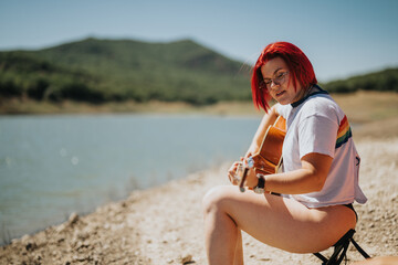 Young lesbian girl with red hair enjoying playing guitar by the lake. Serene outdoor setting, sunny day, and peaceful atmosphere.