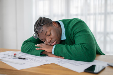 A young black man in a green sweater rests his head on his arms at a desk cluttered with design...