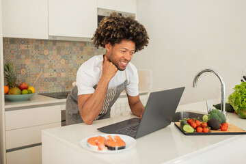 A young black man is focused on a laptop while preparing sushi in a bright kitchen, surrounded by fresh ingredients like vegetables and fish on a counter.