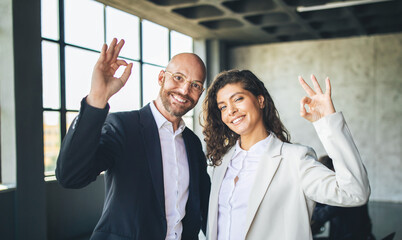 In a modern office with large windows, a man and a woman share a confident smile while making an...