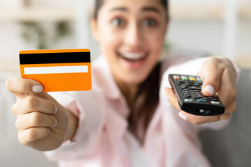 Closeup of amazed excited millennial woman sitting on couch in living room, using tv remote controll and holding yellow golden credit debit card, selective focus on technology, blurred background