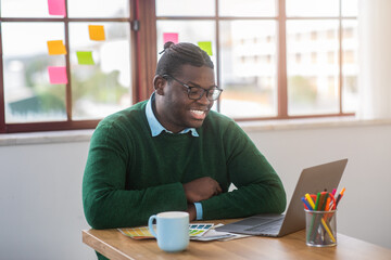 A young black man sits at a wooden table, smiling while using a laptop. Colorful sticky notes are visible on the window behind him, creating a vibrant workspace atmosphere.