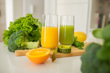 Two glasses filled with vibrant orange and green juices sit on a wooden board. Surrounding them are...