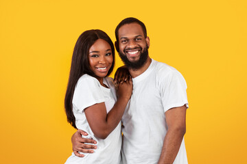 Naklejka premium Happy Loving Family. Portrait of handsome millennial black man embracing his beautiful beloved woman, couple in casual white t-shirts standing and posing isolated over yellow orange studio background