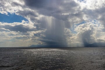 Rainfall over the sea with dramatic clouds, sunlight reflections, and distant mountains on the...