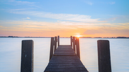 Fototapeta premium Water and lake with wooden jetty (pier) with the setting sun on the horizon and a cloudscape sky in beautiful colors. Countryside landscapes of Netherlands, Giethoorn