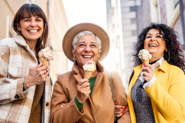 Three mature women eating ice cream cone outside at city urban street - Group of smiling older female friends having fun walking together outside - Family mature and Joyful elderly lifestyle concept