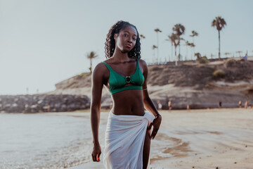 An African woman wearing a green bikini and a white sarong walks on the beach