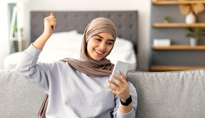 Yes, Great News. Portrait of excited muslim woman in hijab using smartphone gadget, shaking clenched fist, celebrating victory or win, sitting on the comfortable sofa at home, modern interior