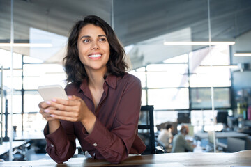 Portrait of young dreaming Indian business woman manager ceo using cell phone mobile app. Pondering Latin Hispanic female businesswoman holding smartphone in office for trading, reading, work online