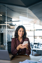 Young Indian business woman manager ceo using cell phone mobile app laptop. Smiling Latin Hispanic mature female businesswoman holding smartphone in office trading, reading, working online. Vertical