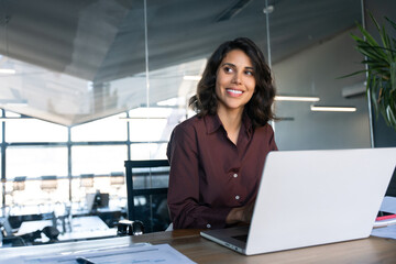 Portrait of smiling latin hispanic middle-aged business woman work on laptop computer in modern office. Indian young businesswoman professional employee using pc, looking dreaming aside. Copy space