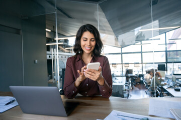 Young Indian business woman manager ceo using cell phone mobile app laptop. Smiling Latin Hispanic mature female businesswoman holding smartphone in office trading, reading, working online. Copy space