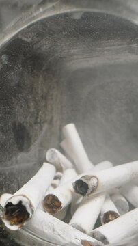 Vertical video. Dolly slider shot moving inside a glass jar filled with cigarette butts, capturing the texture of ashes and used filters in a confined, gritty space, emphasizing discarded items and wa