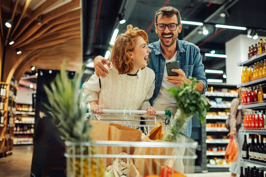 Happy couple using smartphone and pushing shopping cart in supermarket