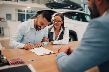 Content couple signing car purchase agreement at dealership table.
