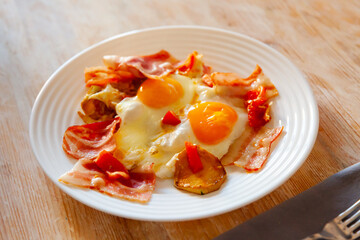 Delicious fried eggs with slices of roasted bacon, potatoes and tomatoes served for breakfast..