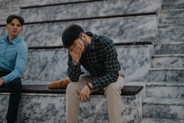 A businessman sits on marble steps, covering his face with one hand, conveying stress and overwhelm. Another man sits nearby, observing the situation.