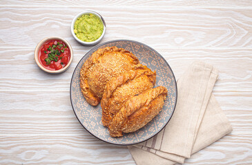 Three freshly baked empanadas with sesame seeds in a bowl, white background, served with tomato salsa and guacamole. Traditional dish of Spain and Latin America