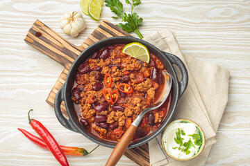 Traditional mexican chili con carne with lime, red chili peppers, and sour cream in a cast iron pan on a wooden board, white background