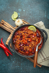Mexican chili con carne served in a cast iron pan garnished with lime wedges and chili pepper slices placed on a wooden cutting board next to fresh chili peppers and garlic