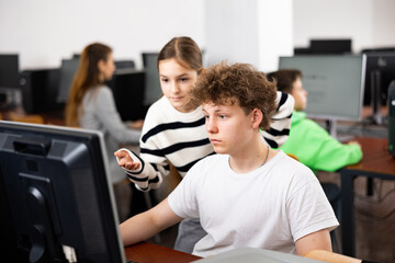 Fototapeta premium Teenage boy sitting at table and helping young girl with PC problem in computer class.