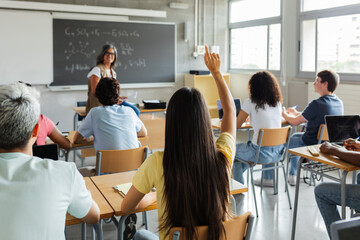 Young group of gen z students attending class at high school. Rear view of female student asking a...