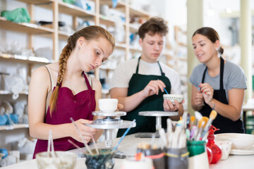 Young woman teacher in apron teaches teenage boy and girl students to make ceramic cup in ceramic workshop