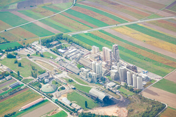 Cement plant in Austria view from above. Concrete plant, surrounded by agricultural fields. Facility includes multiple tall silos, various buildings, and infrastructure such as roads and rail tracks © Leilani