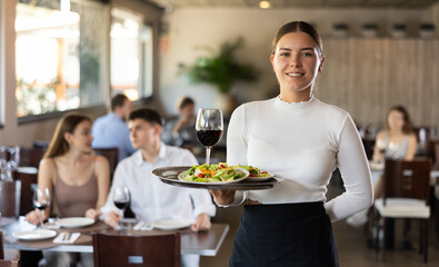 Smiling waitress standing with salad and glass of wine on serving tray in restaurant