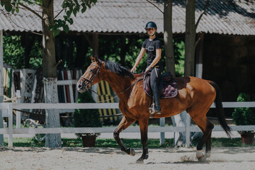 A skilled rider wearing a helmet confidently rides a brown horse in a sunlit outdoor arena,...