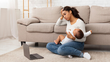 Exhausted Black Mother Holding Crying Baby And Touching Head Suffering From Headache Sitting At Laptop On Floor At Home. Motherhood Burnout Concept. Tired Mother Posing With Shouting Child