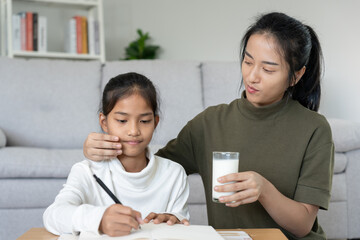 Asian young little girl learn at home. Do homework with kind mother help, encourage for exam. Mom pass on a glass of milk to daughter. Girl happy Homeschool. Mom teach and advice education together.