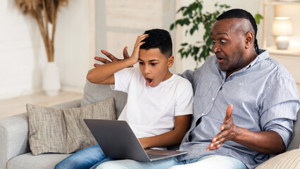 Shock Content. Portrait of mature black man and his grandson looking at laptop screen with open mouth, sitting on sofa at home