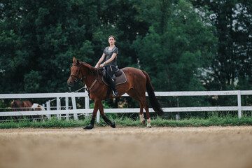 Young woman riding a brown horse in an outdoor equestrian arena surrounded by lush green trees and...