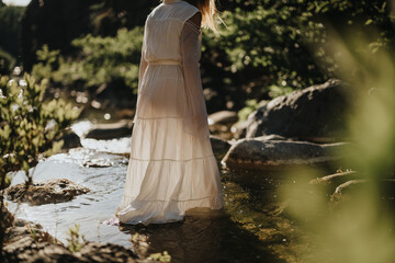 A woman wearing a long white dress stands in a tranquil forest stream, capturing a moment of...