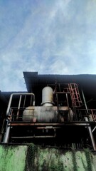 Rusty industrial machinery at an old factory, featuring large cylindrical tanks, pipes, and platforms under a metal roof against a partly cloudy sky.