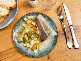 There is plate with fried cod fillet and stewed vegetable on wooden table. Stewed string beans with carrots complement fried fish