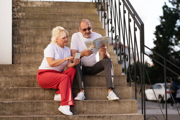 Happy couple engages in a discussion while seated on stone steps, looking at a map together. They are dressed casually and enjoying their time outdoors in a city setting under sunlight.
