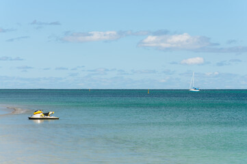 Obraz premium Near Perth, Western Australia: A peaceful coastal scene with a yellow and white jet ski floating in shallow turquoise waters. A sailboat is visible on the horizon beneath a partly cloudy sky.