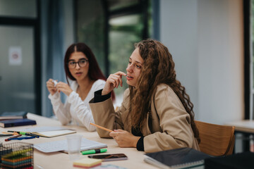 Two young women sitting at a table, discussing educational topics in a bright classroom setting, with study materials spread around them.