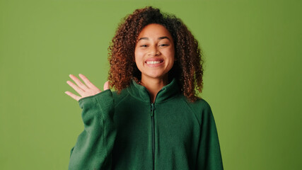 Young woman wearing casual clothes waving her hand and smiling to camera on green screen background in the studio. Positive woman shows a gesture of greeting, happy to see, saying hello and waving © Andrii Nekrasov